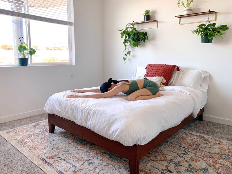 Person in green workout set kneeling in child's pose on a white bed in a bright bedroom with hanging plants and a red pillow.