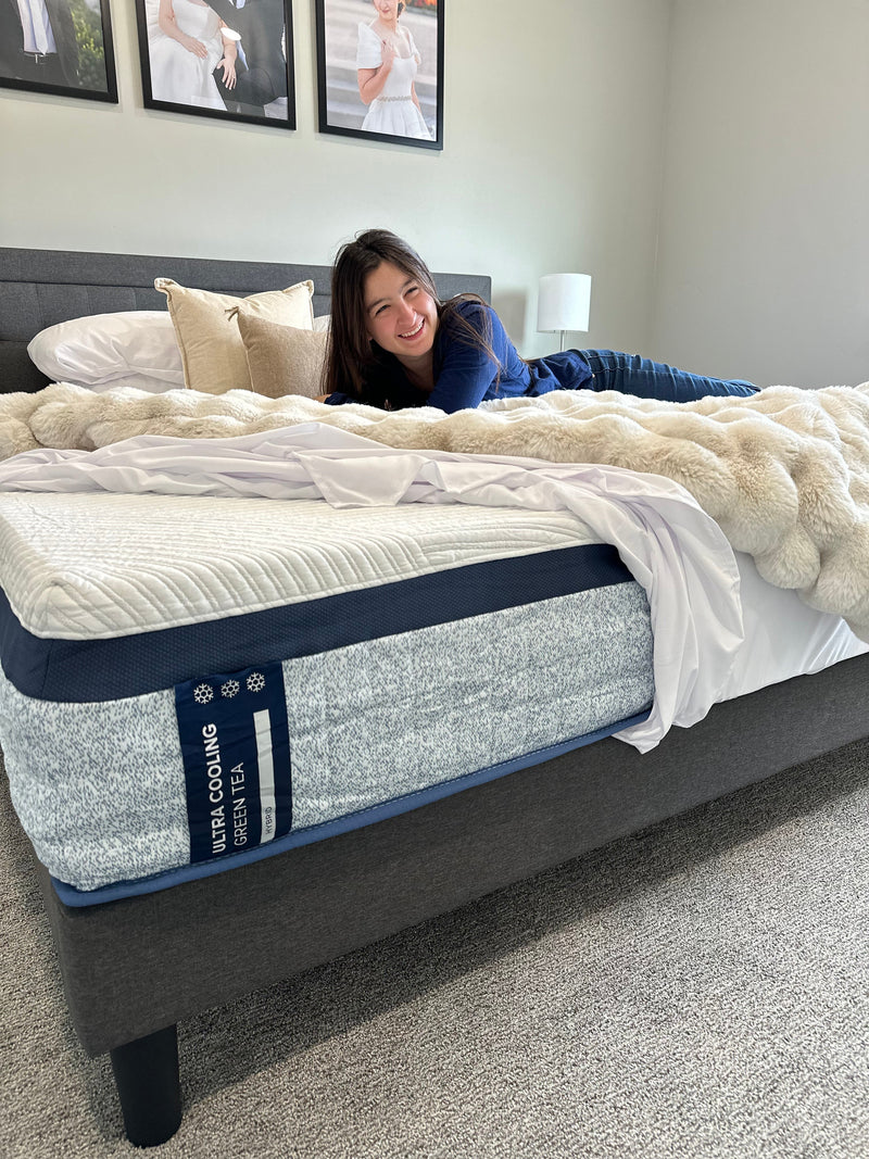 Person smiling while lying on a bed showcasing an Ultra Cooling Green Tea mattress with a textured white top and blue cooling label, highlighting comfort and the mattress’s cooling support in a bright bedroom