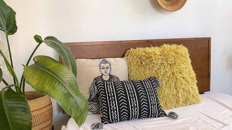 Cozy bed with a wooden headboard, decorated with a yellow shag pillow, a black patterned pillow with tassels, and a beige pillow featuring a Buddha illustration, next to large green houseplants and woven baskets for a warm, earthy look.