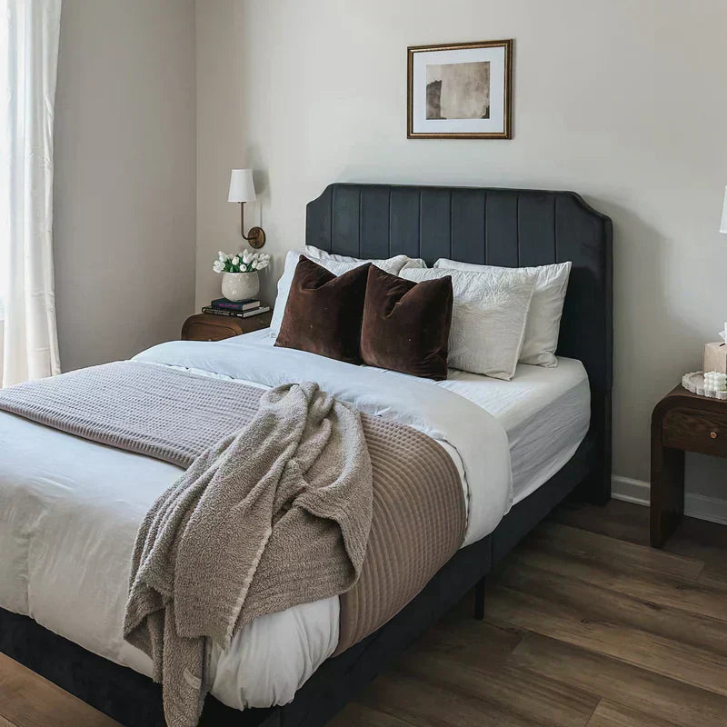 Upholstered bed with dark paneled headboard styled with layered white bedding, brown accent pillows, and a textured taupe throw in a bright, modern bedroom, emphasizing a cozy setup with wood nightstands and soft natural light.