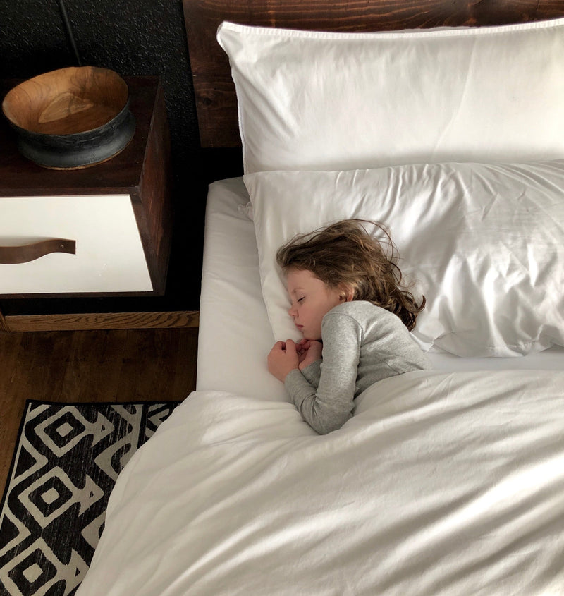 Child sleeping on a bed with white bedding in a cozy room, shown beside a wood nightstand and patterned rug to highlight a peaceful sleep environment.