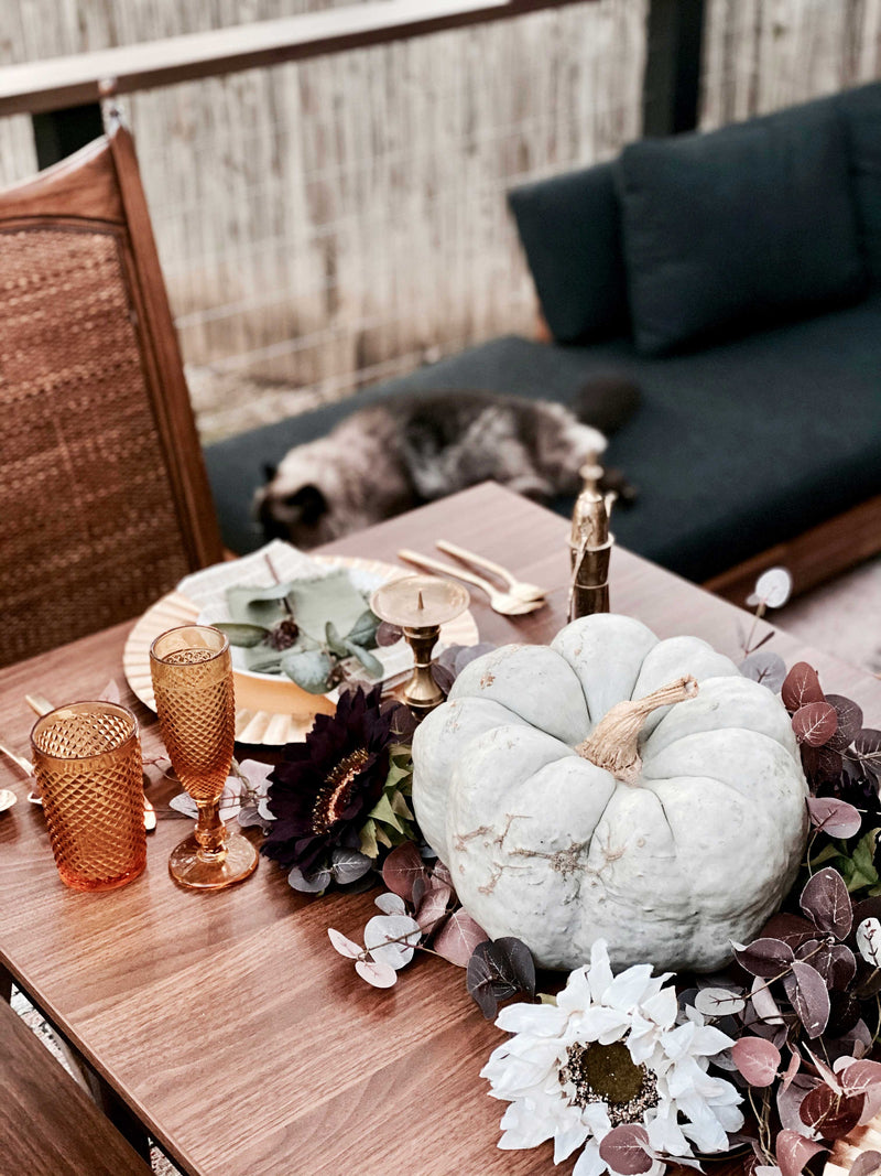 Fall-themed outdoor table with amber glasses, brass accents, and a pale pumpkin centerpiece surrounded by flowers, with a cat resting on a dark cushioned bench in the background