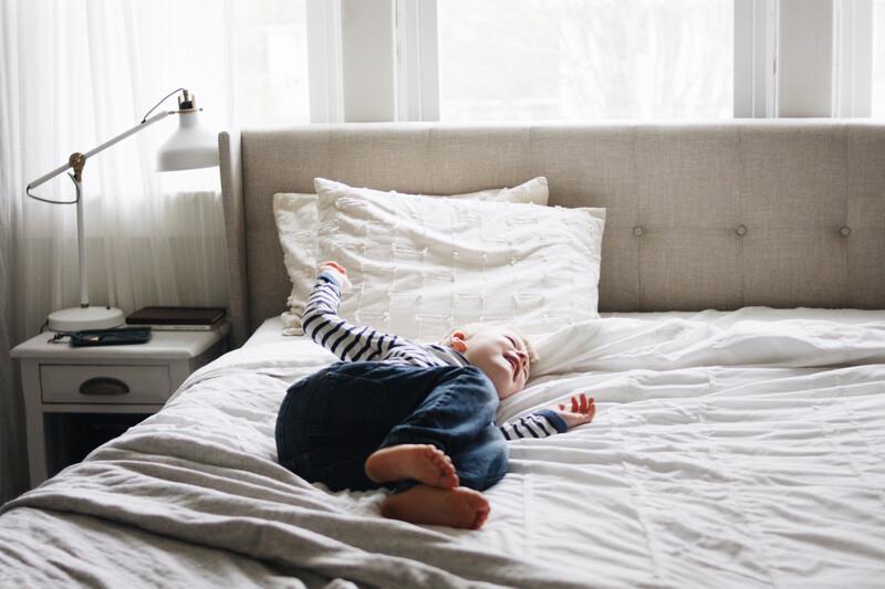 Zinus Soft upholstered bed with white bedding in a calm, airy bedroom; a toddler rests comfortably on the mattress, conveying gentle support, everyday comfort, and a peaceful sleep environment designed for family-friendly use.
