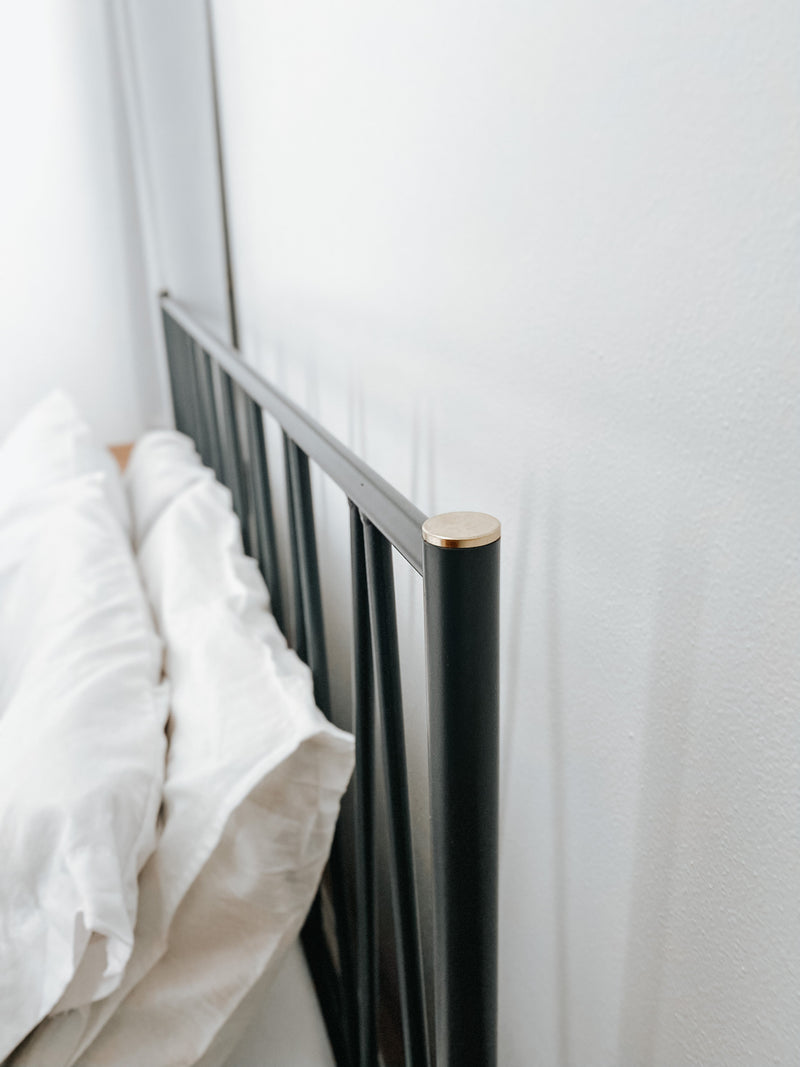 Close-up of black metal bed frame headboard with slim vertical spindles and a rounded wood-accent cap, shown beside white pillows against a light wall to emphasize sturdy construction, modern design, and smooth finish details