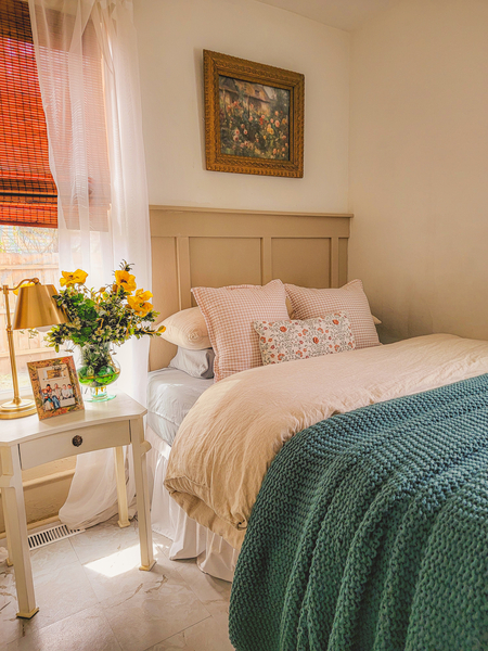 Warm, sunlit bedroom with layered neutral bedding, floral pillow, and flowers on a bedside table