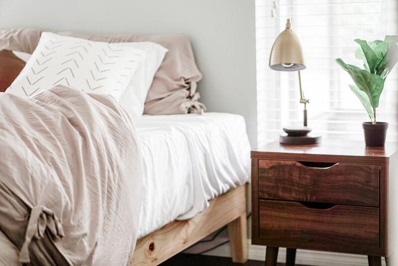 Close view of bed with soft neutral bedding and a wood nightstand holding a lamp and plant.