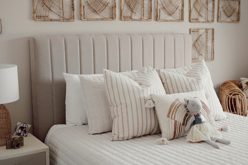 Upholstered bed with a vertically channeled beige headboard, layered white and striped pillows, and a textured quilt, styled with woven wall decor and a plush toy to highlight soft materials, relaxed comfort, and a calm, family-friendly bedroom setting