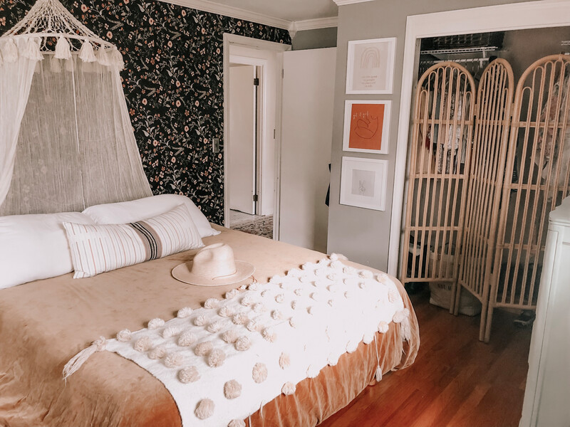 Boho-style bedroom with upholstered bed dressed in warm tan bedding and textured white throw, styled with patterned wallpaper, macramé canopy, and layered pillows to highlight a cozy, eclectic sleep space with inviting comfort and natural accents
