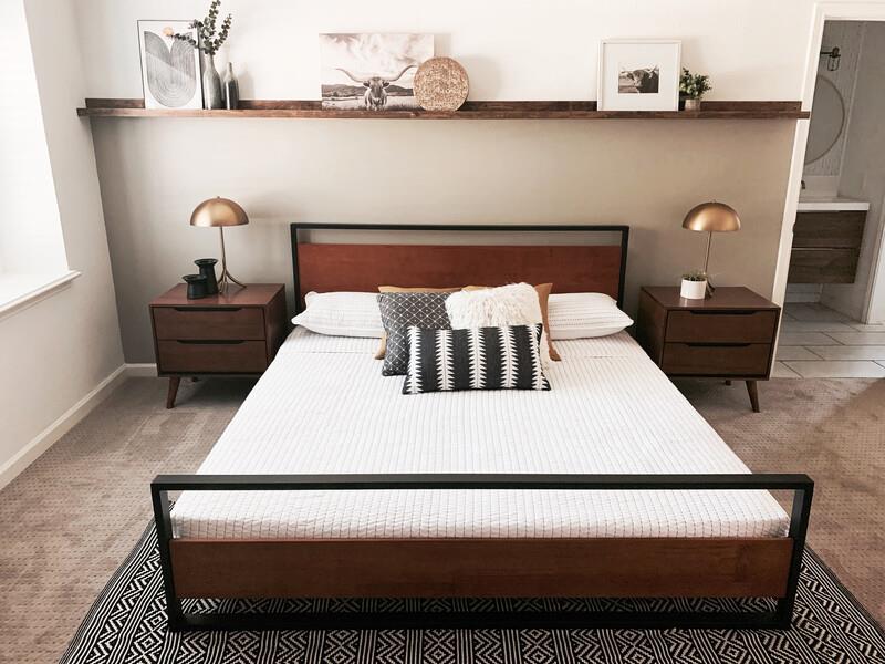 Mid-century modern platform bed with a wood headboard and black metal frame, dressed in white bedding and patterned accent pillows; styled with matching wood nightstands and brass dome lamps