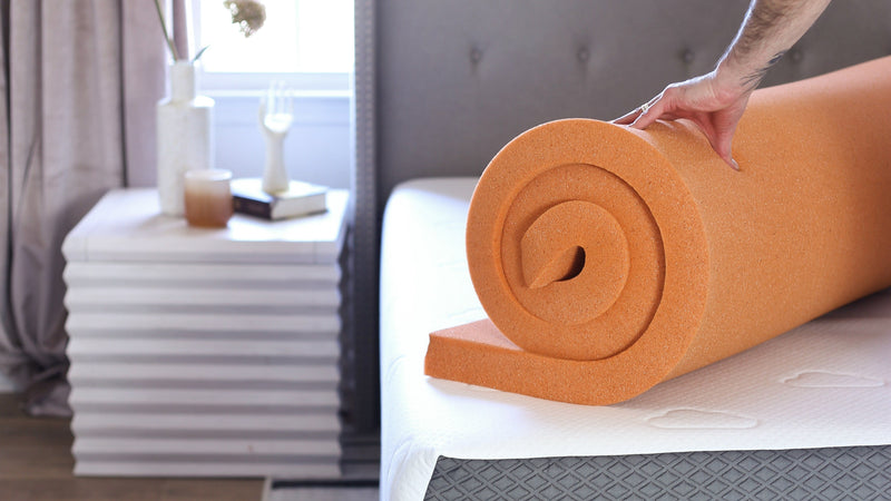 Hand unrolling an orange foam mattress topper onto a bed, showing flexible material and easy setup in a bright bedroom with simple decor.