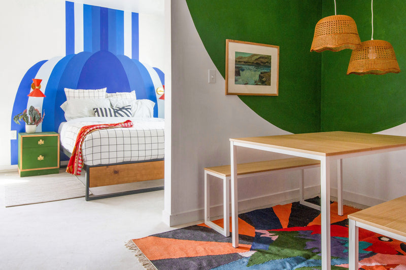 Colorful home space with a vibrant blue-and-white mural behind a modern bed and an adjacent dining area featuring green walls, woven pendant lights, and a bright patterned rug.