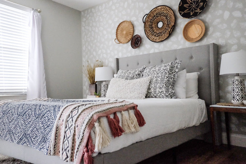 Cozy bedroom with a gray tufted bed, patterned pillows, tassel blanket, and woven baskets displayed on a textured accent wall.