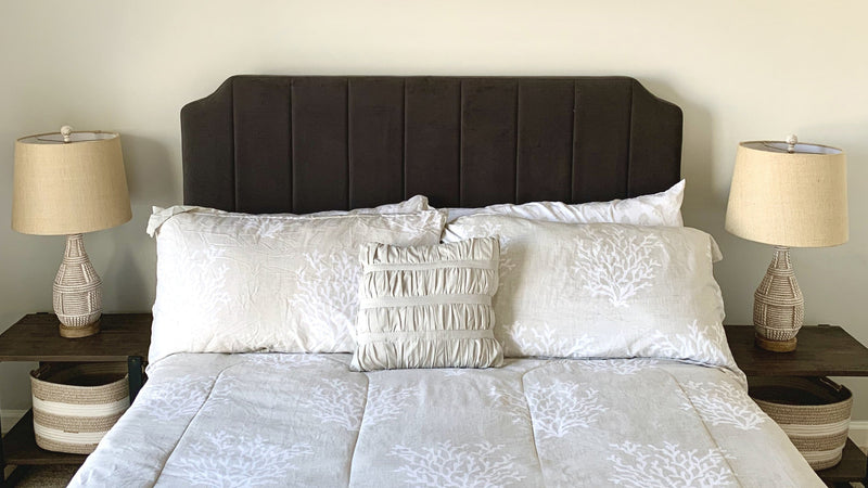 Neutral bedroom with a dark upholstered headboard, light patterned bedding, and matching woven lamps on wooden nightstands