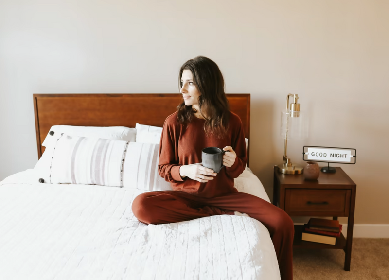 Person sitting cross-legged on a neatly made bed with a wooden headboard, holding a mug and looking to the side, styled with white bedding, striped pillows, and a bedside table featuring a lamp, books, and a small sign that reads “Good Night.