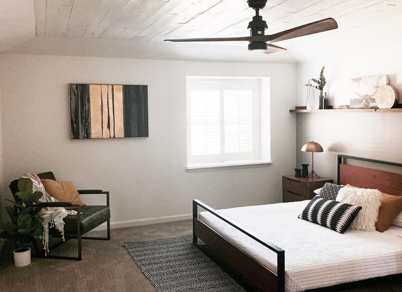 Neutral bedroom with a wood-and-metal bed frame, textured bedding, a dark rug, and a reading corner, styled with warm accents and a ceiling fan for a calm, modern look.