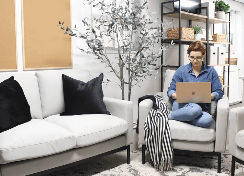 Modern office space with white armchairs, black pillows, shelving with baskets, and a person working on a laptop.