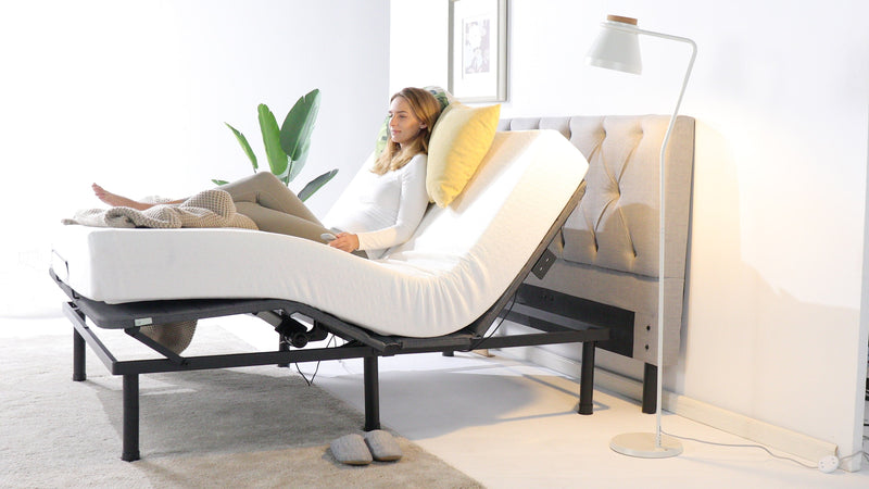 Person relaxing on an adjustable bed with the head and legs raised, featuring a supportive mattress, soft bedding, and a modern setup with a tall lamp and potted plant.