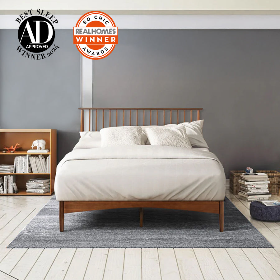 Modern bedroom with wood bed frame and neutral bedding against gray wall, styled with bookshelves, pillows, and woven basket; badges include AD Best Sleep Winner 2024 and Real Homes So Chic Awards Winner, highlighting recognition for sleep comfort and design. 