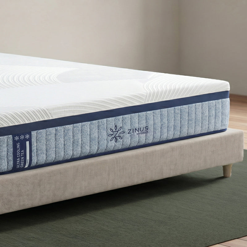 Zinus Balanced Comfort Luxe Hybrid Mattress, close-up corner view showing the quilted white top cover with wave-pattern stitching, navy trim, and channel-quilted light blue woven side panel with Zinus Hybrid brand tag and triple cooling icons, on an upholstered bed.