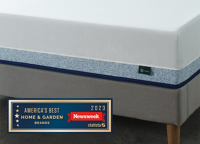 Close-up of Zinus mattress with white top and blue patterned side panel on gray upholstered bed, featuring Newsweek 2023 America's Best Home & Garden Brands award badge.