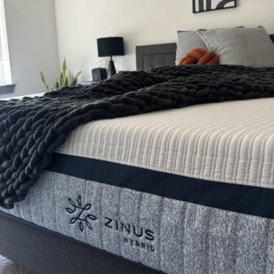 Zinus Ultra Cooling Hybrid mattress with quilted white top and gray side panel labeled Hybrid, styled with a chunky black knit throw and neutral decor, emphasizing responsive coil support, pressure-relieving comfort, and a modern bedroom aesthetic.