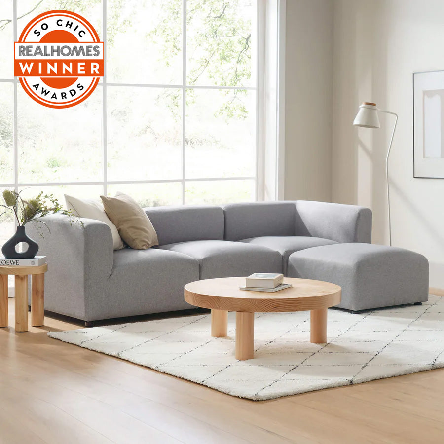 Modern living room with large window, light wood floors, and minimalist decor featuring gray sectional sofa with chaise, round wood coffee table, white rug with diamond pattern, side table with books and vase, and floor lamp; badge reads So Chic Real Homes Awards Winner 