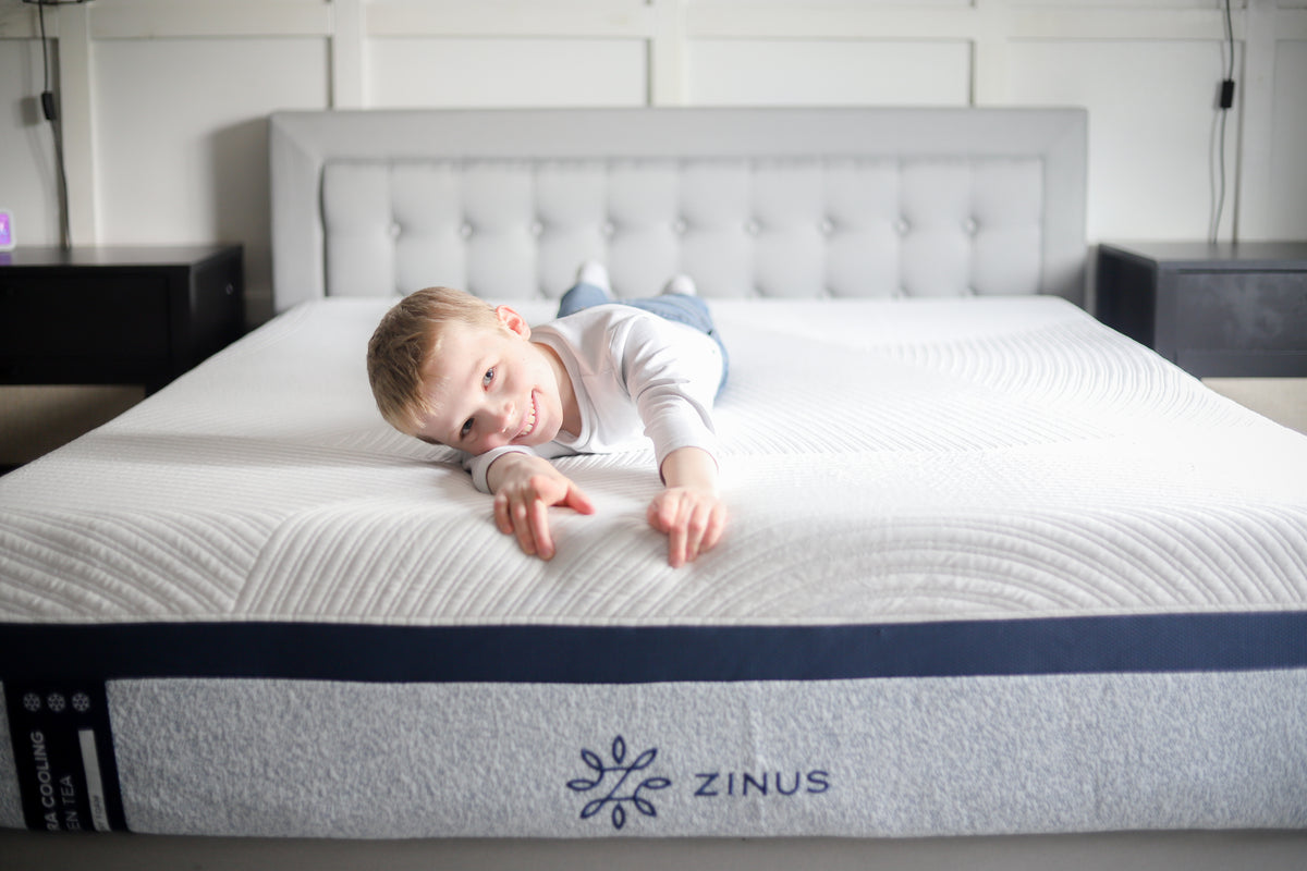 Smiling child lies across a Zinus Ultra Cooling Green Tea Hybrid mattress with white quilted top and gray-blue side panel, styled on an upholstered bed frame with tufted gray headboard in a bright modern bedroom, highlighting comfort and playful relaxation