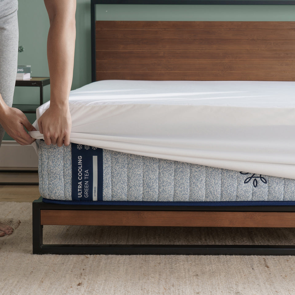 Person tucking white sheet over Zinus Ultra Cooling Green Tea Hybrid mattress with blue patterned side panel, shown on modern wood and metal platform bed in a cozy bedroom
