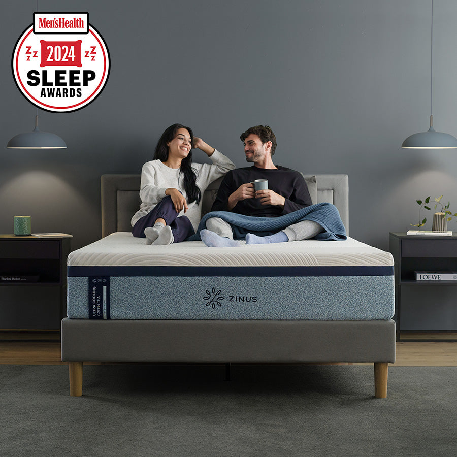 Men’s Health 2024 Sleep Awards badge with couple sitting on Zinus Ultra Cooling Green Tea Hybrid mattress in a modern bedroom, smiling and relaxing with mugs and blanket, highlighting recognition for supportive comfort and award-winning sleep innovation. 