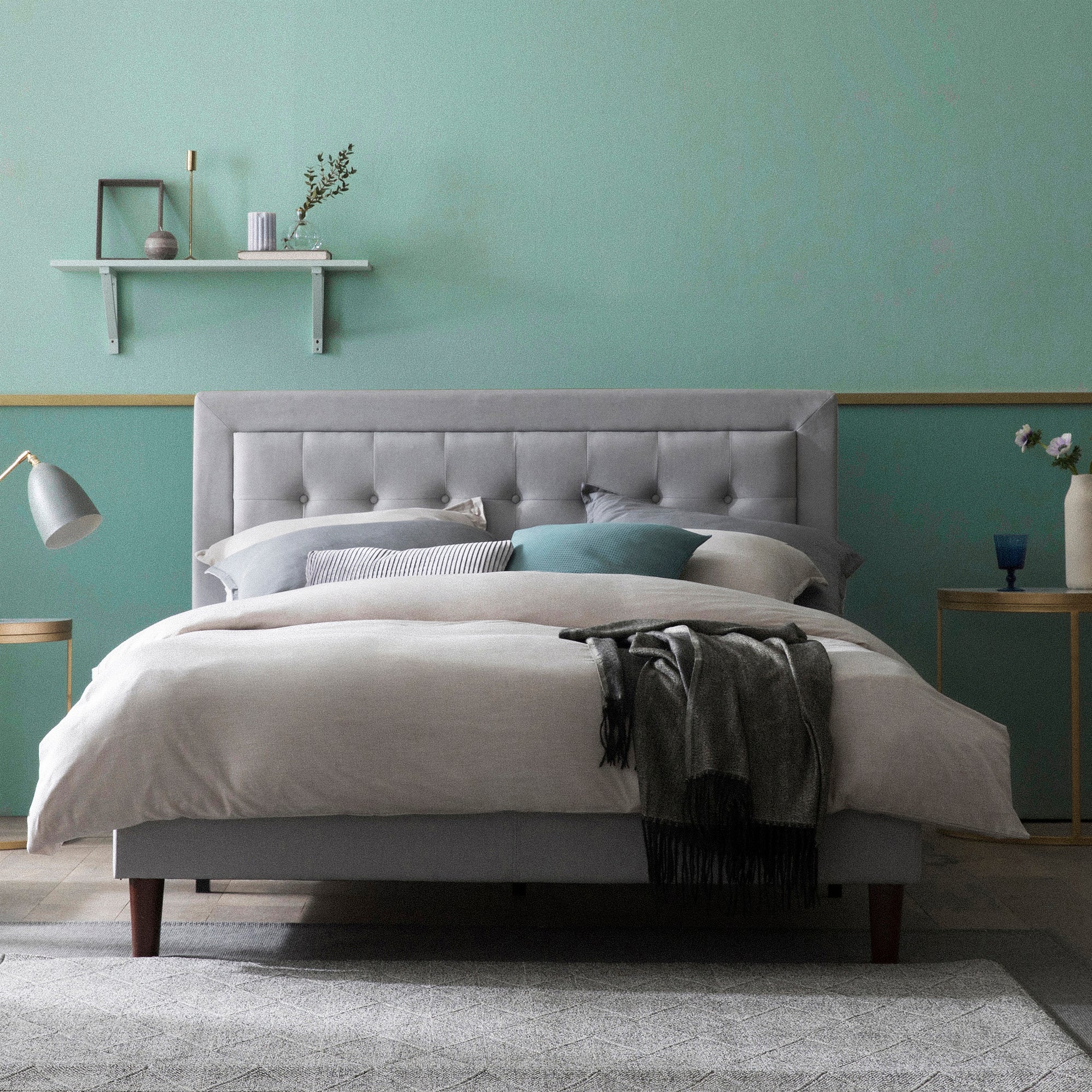 Gray upholstered platform bed with tufted headboard styled in a modern bedroom, featuring neutral bedding, accent pillows in teal and gray, and a dark throw blanket; room includes a round side table with lamp, chair, and decorative shelf against a two-tone green wall.