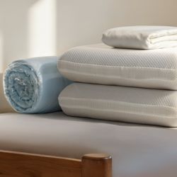 Stack of Zinus bedding essentials including folded white mattress toppers, rolled light blue comforter, and folded white sheets, placed neatly on a wooden bed frame in a sunlit room, highlighting layered sleep accessories for added comfort and support.