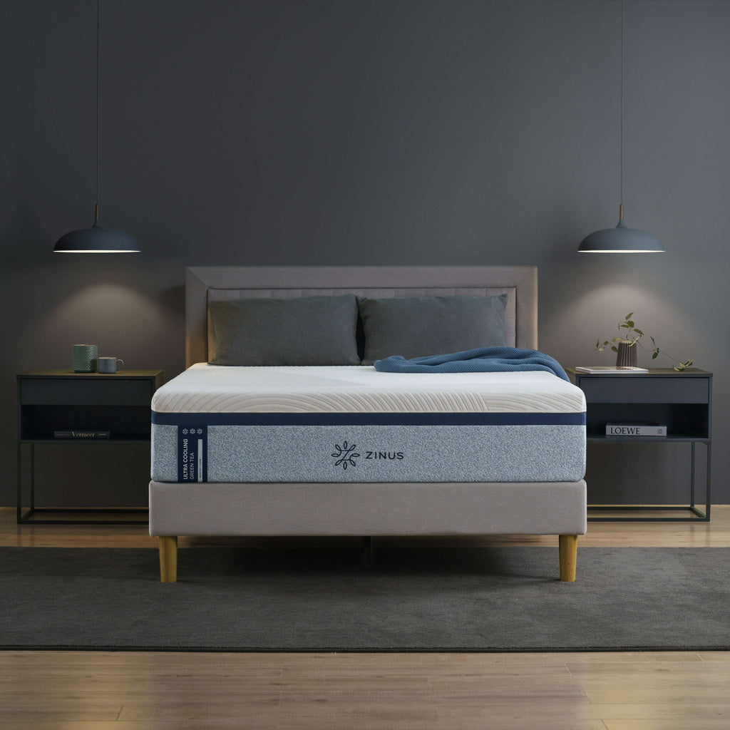 Zinus Ultra Cooling Green Tea Memory Foam Mattress styled in a modern bedroom with gray upholstered bed frame and wood legs. The mattress features blue accent side panels and a smooth cooling cover, designed to regulate temperature, ease pressure points, and support deep sleep 
