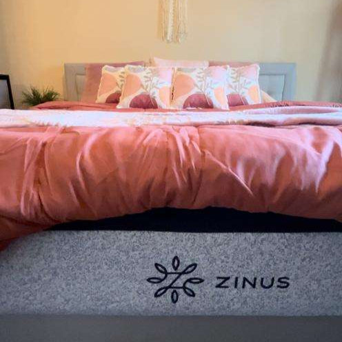 Zinus memory foam mattress with gray fabric cover and visible logo, styled on a modern bed with coral pink bedding and patterned pillows, highlighting supportive comfort and clean design suited for a cozy, contemporary bedroom setting.