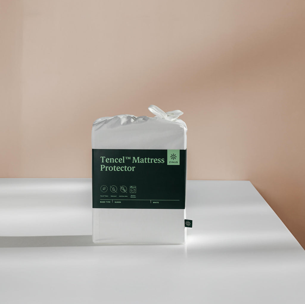 Packaged Zinus TENCEL waterproof mattress protector in white shown on a table, featuring dark green label with brand logo and icons highlighting breathable fabric, water resistance, and machine washability. Protector provides protective comfort and extends mattress longevity with ease. 