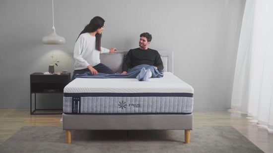 A couple sits on a bed, engaging in conversation. The room features a modern design with a bedside table and a lamp.